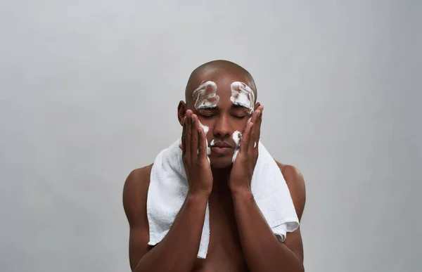 Shirtless joven afroamericano hombre lavándose la cara con limpiador de espuma facial mientras posa con toalla alrededor de su cuello aislado sobre fondo gris — Foto de Stock