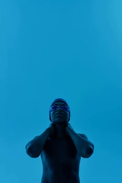 Futuro campeón. Adolescente africano niño en gorra de natación y gafas manteniendo los brazos en su cuello y mirando al revés mientras posando aislado sobre fondo azul —  Fotos de Stock