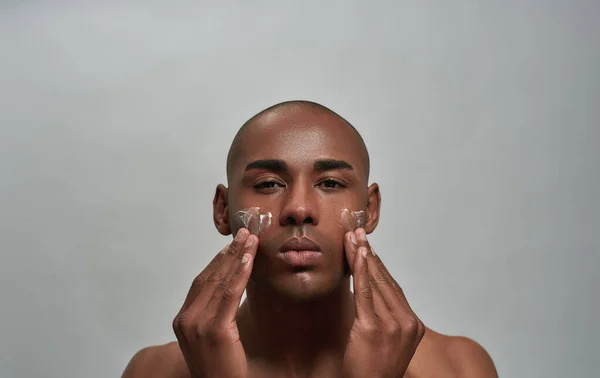 Retrato de un joven afroamericano guapo mirando a la cámara, aplicándose crema en la cara, posando aislado sobre un fondo gris — Foto de Stock