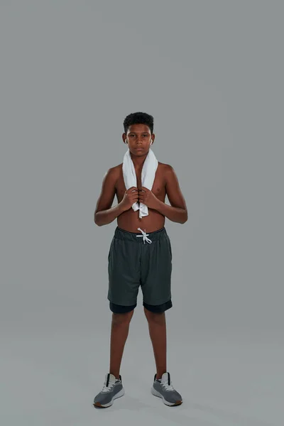 Tiro vertical de um adolescente africano semi-nu com toalha nos ombros usando fones de ouvido sem fio olhando para a câmera enquanto estava contra fundo cinza — Fotografia de Stock