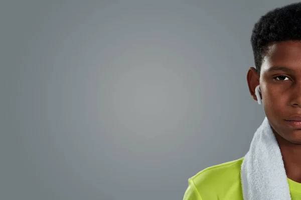 Portrait demi-visage d'un adolescent africain confiant avec une serviette blanche sur les épaules portant des écouteurs sans fil regardant la caméra tout en posant sur fond gris — Photo