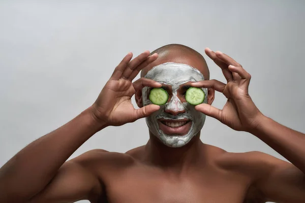 Porträtt av positiva unga afrikanska amerikanska mannen ler mot kameran, håller gurka skivor medan du använder ansiktsmask, poserar isolerad över grå bakgrund — Stockfoto