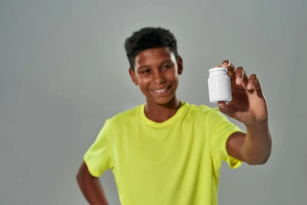 Remaja afrika ceria yang muncul di kamera wadah putih dengan obat-obatan medis, vitamin atau suplemen sambil berdiri melawan latar belakang abu-abu — Stok Foto