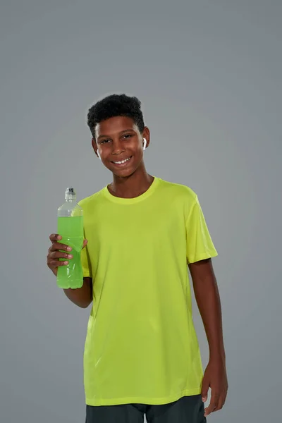 Foto vertical de un alegre adolescente africano vestido con ropa deportiva sosteniendo bebida energética, sonriendo a la cámara mientras está de pie sobre un fondo gris —  Fotos de Stock