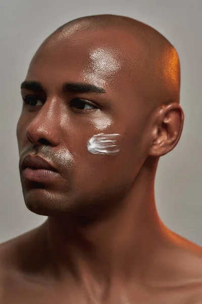 Κοντινό πορτραίτο ενός όμορφου νεαρού Αφροαμερικανού με κρέμα στο μάγουλό του να κοιτάζει αλλού ενώ ποζάρει απομονωμένος σε γκρι φόντο — Φωτογραφία Αρχείου