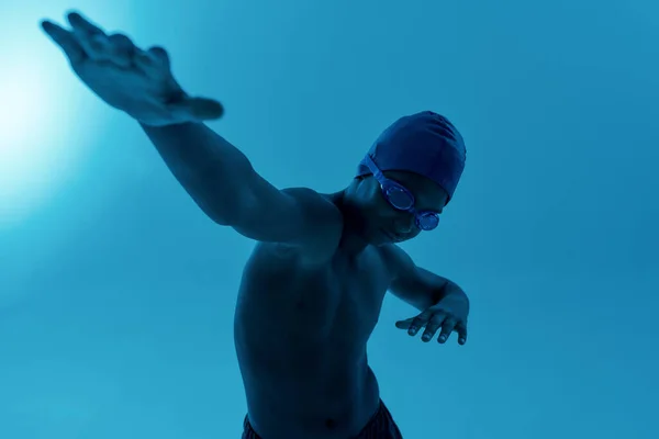 Adolescente Africano menino desportista em touca de natação e óculos tendo treinamento de natação enquanto em pé isolado sobre fundo azul — Fotografia de Stock