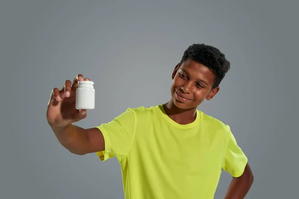 Vitaminpräparate für Jugendliche. Glücklicher afrikanischer Teenager mit weißem Behälter, der ihn ansieht und lächelt, während er vor grauem Hintergrund steht — Stockfoto