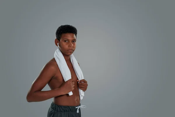 Merasa lelah. Setengah telanjang Afrika anak laki-laki dengan handuk di bahu mengenakan earphone nirkabel menatap kamera sambil berdiri melawan latar belakang abu-abu — Stok Foto