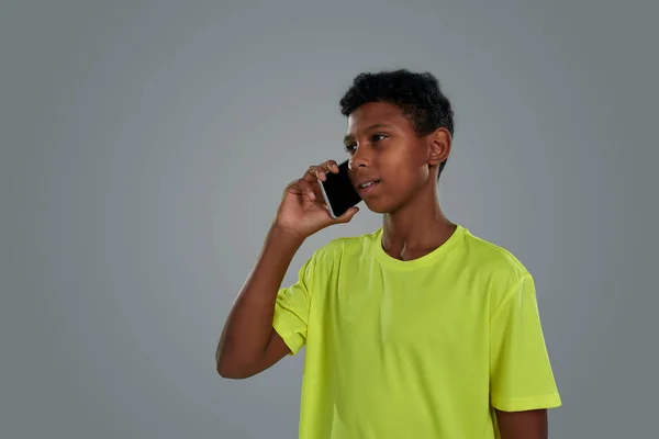 Ungdomar och kommunikationskoncept. Närbild bild av en tonåring afrikansk pojke bär neon t skjorta talar per telefon när du står mot grå bakgrund — Stockfoto