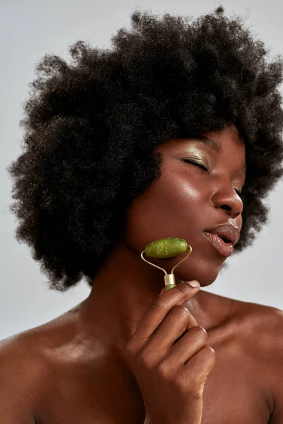 Retrato de cerca de una atractiva joven afroamericana con una piel brillante perfecta usando un rodillo de jade o una herramienta de masaje natural, posando aislada sobre un fondo gris — Foto de Stock