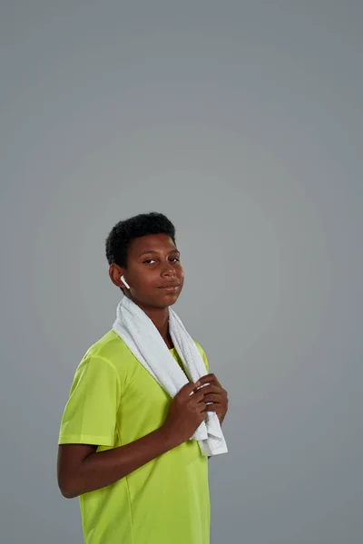 Готовий до тренування. Впевнений підліток спортивний африканський хлопчик в неоновому сорочці з білим рушником на плечах в бездротових навушниках дивиться на камеру — стокове фото