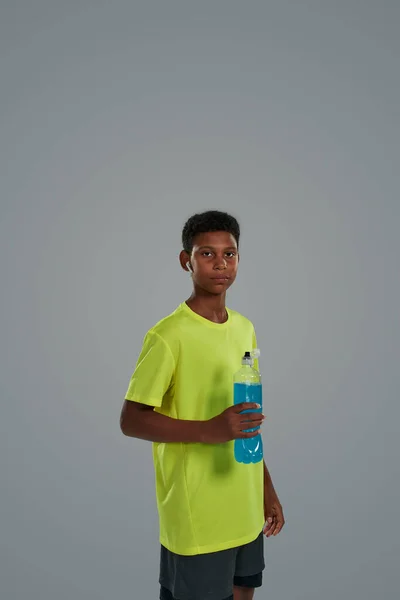 Fotografía vertical de un adolescente deportivo africano sosteniendo una bebida de energía azul mientras está de pie sobre un fondo gris, mirando a la cámara —  Fotos de Stock