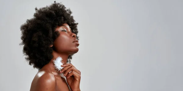 Portret van sensuele Afro-Amerikaanse vrouw met afrohaar die haar gloeiende huid aanraakt met witte veren, poserend met gesloten ogen geïsoleerd over grijze achtergrond — Stockfoto