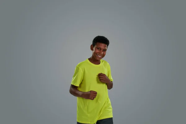 Feliz adolescente africano usando camiseta de neón manteniendo las manos en los puños y sonriendo a la cámara mientras está de pie sobre fondo gris — Foto de Stock