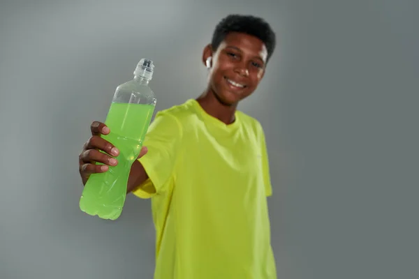 Освіжається під час тренування. Веселий підліток африканський хлопчик в спортивному одязі, що тримає енергетичний напій, посміхається на камеру, стоячи на сірому фоні — стокове фото