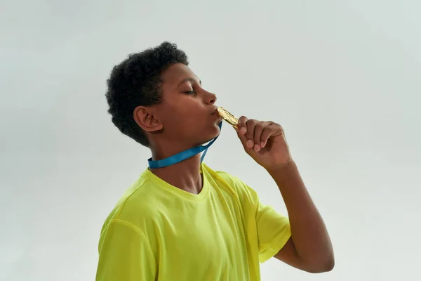 Jovem vencedor. Vista lateral de um menino africano adolescente beijando medalha de ouro enquanto estava isolado sobre fundo cinza — Fotografia de Stock