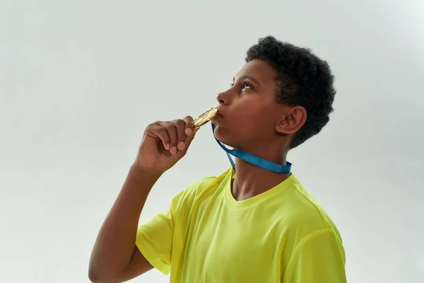 Вид збоку підлітка африканський хлопчик цілує золоту медаль, стоячи ізольовано на сірому фоні — стокове фото