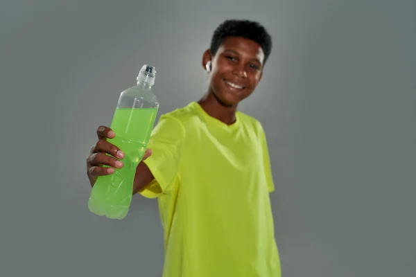 Try it. Cheerful teenage african boy wearing sports clothes showing energy drink at camera and smiling while standing against grey background — Stock Photo, Image