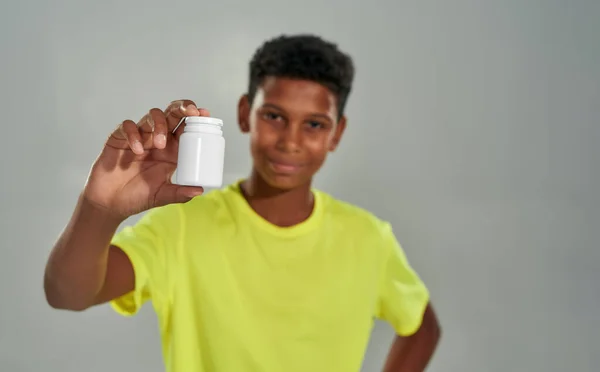Smiling african remaja laki-laki membawa wadah putih dengan obat-obatan selama promosi di kamera sambil berdiri melawan latar belakang abu-abu di studio — Stok Foto