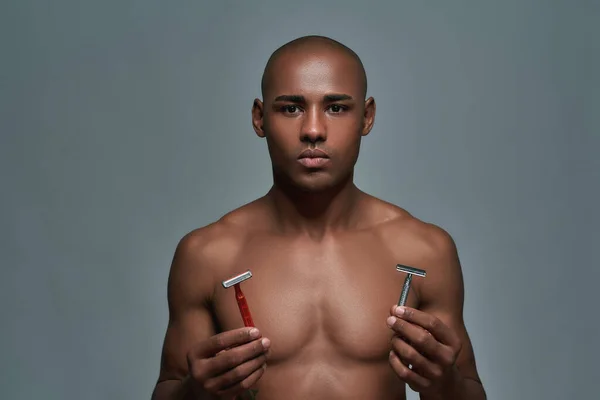Shirtless young african american man looking at camera, choosing between two different types of shaving razors isolated over grey background — Stock Photo, Image