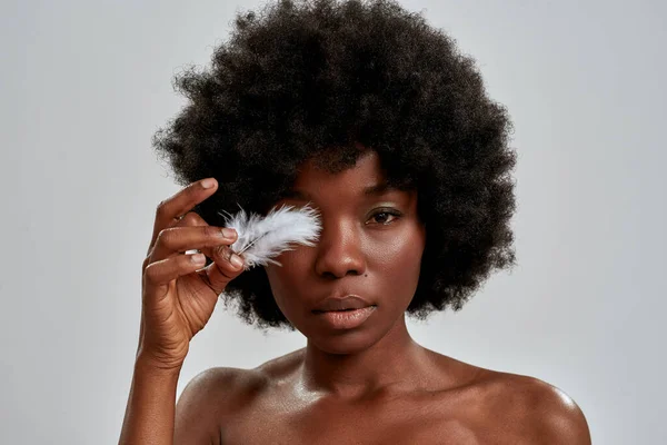 Πορτρέτο του όμορφου αφροαμερικάνικου γυναικείου μοντέλου που κοιτάζει την κάμερα, καλύπτει το ένα μάτι με λευκό φτερό, ποζάρει απομονωμένο πάνω από γκρι φόντο — Φωτογραφία Αρχείου