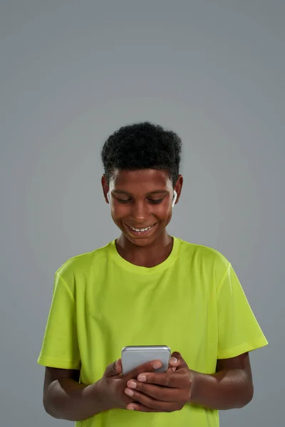 네온 티셔츠를 입고 회색 배경을 등지고 서서 스마트폰을 사용하는 10 대 아프리카 소년에게 미소짓는 모습 — 스톡 사진