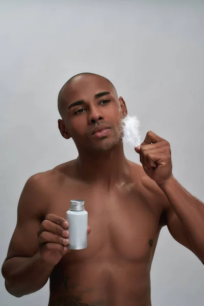 Shirtloser, gutaussehender afrikanisch-amerikanischer Typ, der Rasierwasser in der Hand hält, seine Gesichtshaut mit weißer Feder berührt, isoliert vor grauem Hintergrund posiert — Stockfoto