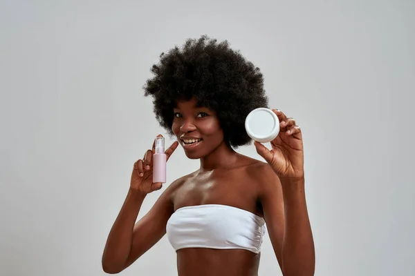 Joyful young african american woman with afro hair smiling at camera, choosing between two skincare products while standing isolated over gray background