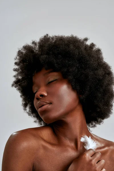 Portret van een mooie Afrikaans-Amerikaanse jonge vrouw met afrohaar die haar huid aanraakt met witte veren, poserend met gesloten ogen geïsoleerd over grijze achtergrond — Stockfoto