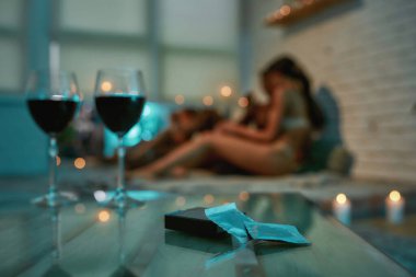 Masada iki kadeh kırmızı şarap ve prezervatif var. Yatakta sevişmeden önce arka planda öpüşen ve sarılan genç yarı çıplak erkek ve kadın var.