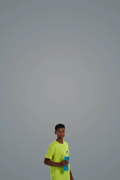 Scatto verticale di un adolescente africano sportivo che tiene in mano una bevanda energetica blu mentre si trova sullo sfondo grigio, guardando la fotocamera — Foto Stock