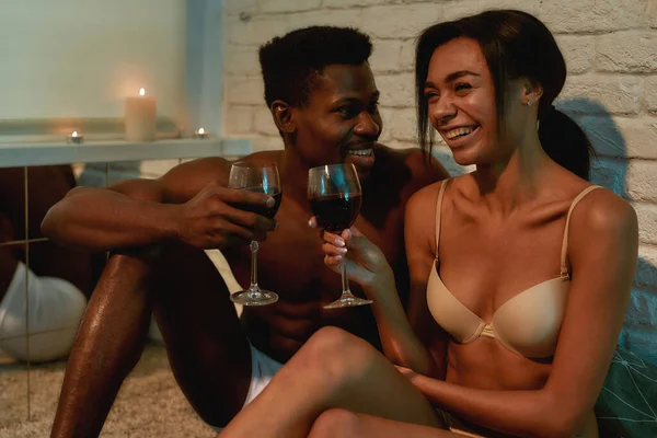 Tineri cuplu african fericit, jumătate bărbat gol și femeie care beau vin, vorbesc și râd în timp ce stau pe pat acasă — Fotografie, imagine de stoc