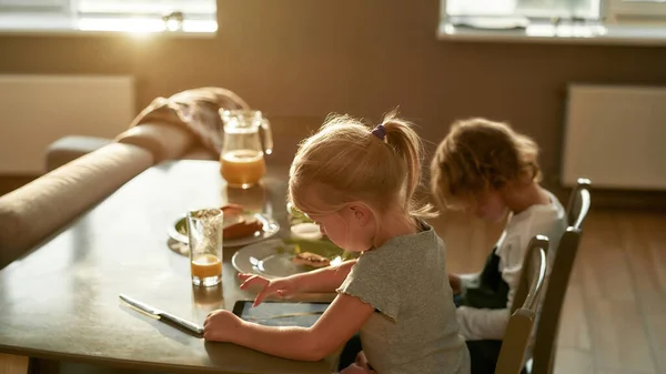 Lindos hermanitos usando dispositivos digitales mientras desayunan o almuerzan, sentados juntos en la mesa de la cocina —  Fotos de Stock