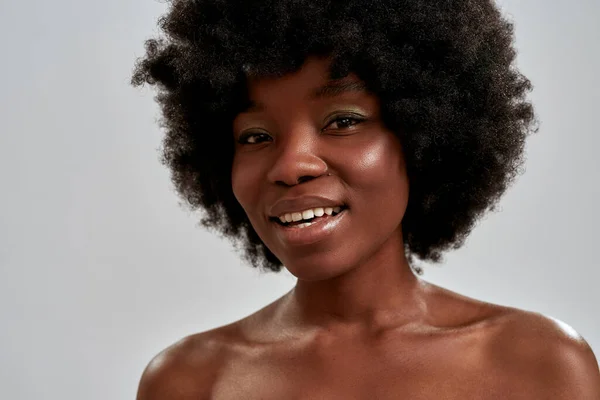 Close-up portret van vrolijke Afrikaanse amerikaanse jonge vrouw met afro haar en perfect glad gloeiende huid glimlachen op camera terwijl poseren geïsoleerd over grijze achtergrond — Stockfoto