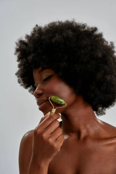 Közelkép portré nyugodt fiatal afro-amerikai nő tökéletes bőr segítségével jáde görgő vagy természetes masszázs eszköz, pózol elszigetelt szürke háttér — Stock Fotó