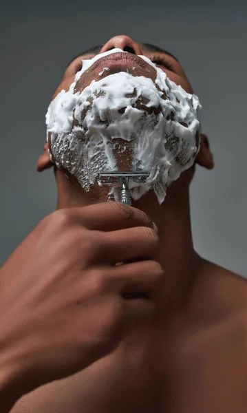 Dekat pemuda african american pria dengan busa diterapkan di wajahnya menggunakan pisau cukur baja untuk menghapus jerami dari leher terisolasi atas latar belakang abu-abu — Stok Foto