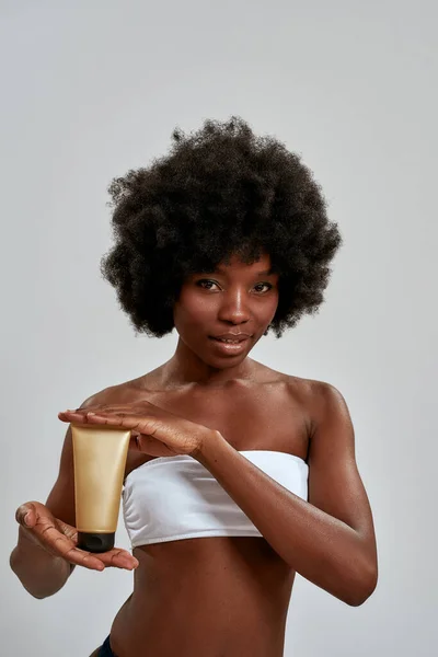 Bastante joven mitad desnudo mujer afroamericana sosteniendo botella de loción corporal y mirando a la cámara, posando aislado sobre fondo gris — Foto de Stock