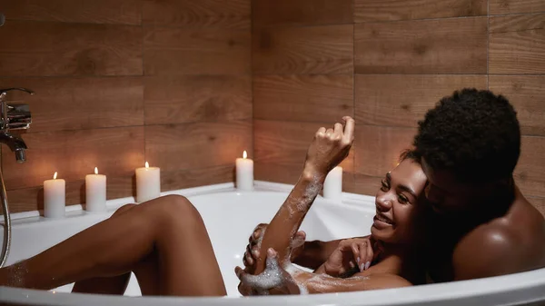 Happy with you. Young beautiful heterosexual african couple, man and woman taking foamy bath with candles together, enjoying romantic evening at home