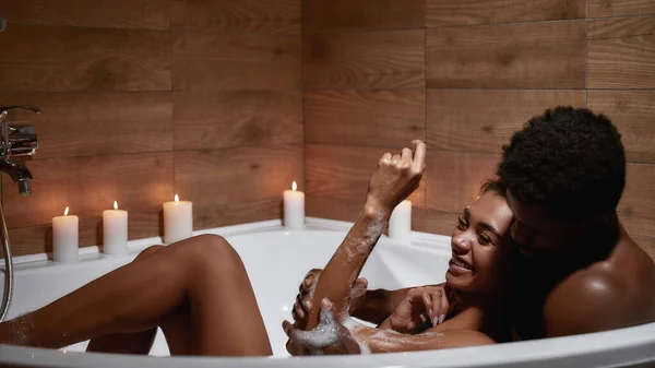 Feliz contigo. Joven hermosa pareja heterosexual africana, hombre y mujer tomando un baño espumoso con velas juntos, disfrutando de una velada romántica en casa —  Fotos de Stock