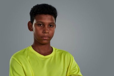 Gri arka plana karşı poz verirken kameraya bakan neon tişört giyen özgüvenli Afrikalı bir çocuğun portresi.