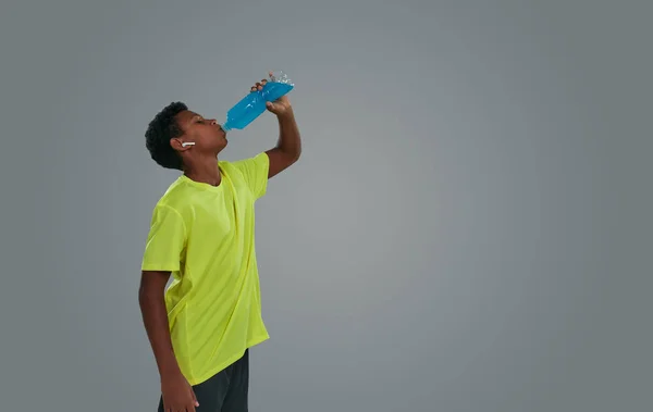 Kablosuz kulaklık takan, gri arka planda dikilirken mavi enerji içeceği içen Afrikalı bir çocuğun yan görüntüsü. — Stok fotoğraf