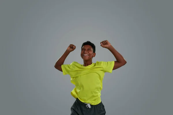 Emocionado adolescente africano con camiseta de neón celebrando el éxito, manteniendo los brazos levantados mientras está de pie sobre un fondo gris — Foto de Stock