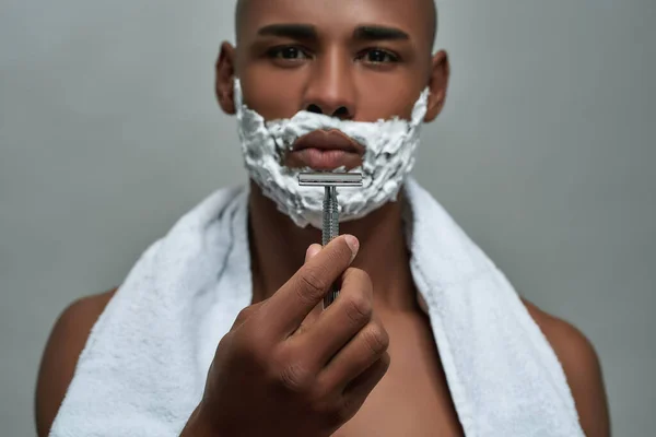 Potret pemuda tampan african american pria dengan busa cukur diterapkan di wajahnya menatap kamera, memegang pisau cukur baja, berpose terisolasi atas latar belakang abu-abu — Stok Foto