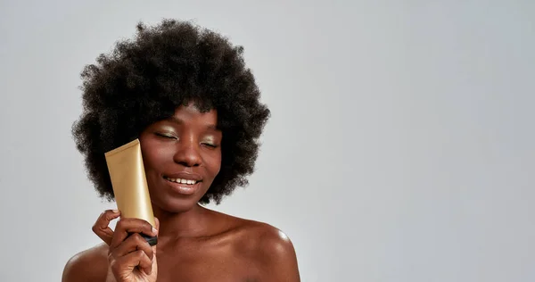 Güzel, çıplak, Afro-Amerikan bir kadının vücut losyonu şişesini tutarken güzel portresi ve gözleri kapalı gri arka planda poz verirken gülümsemesi. — Stok fotoğraf