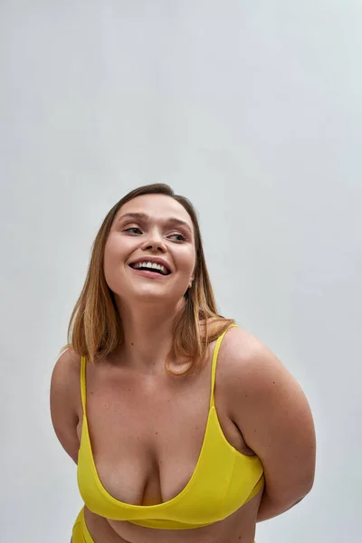 Mujer joven caucásica con sujetador amarillo sonriendo a un lado, posando aislado sobre fondo gris —  Fotos de Stock