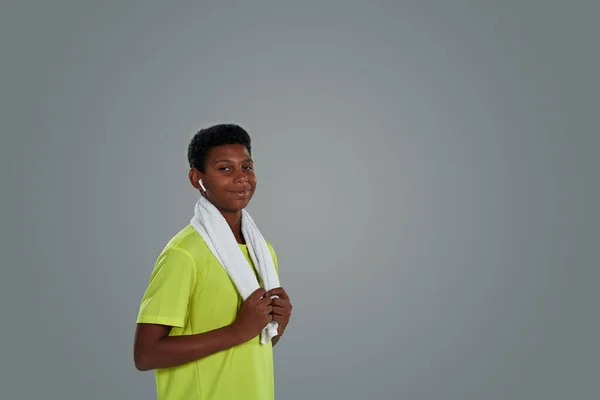Retrato de um adolescente feliz menino africano esportivo em camisa de néon com toalha branca nos ombros usando fones de ouvido sem fio olhando para a câmera enquanto posando contra fundo cinza — Fotografia de Stock