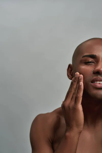 Half gezicht portret van shirtloze jonge Afro-Amerikaanse man aanbrengen lotion op zijn gezicht, poseren geïsoleerd over grijze achtergrond — Stockfoto