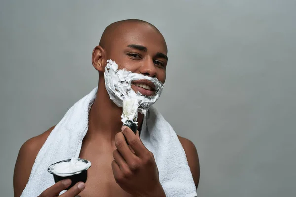 Pemuda african Amerika yang tidak menarik tersenyum pada kamera, menggunakan kuas sambil mengenakan busa cukur di wajahnya, berpose terisolasi di atas latar belakang abu-abu — Stok Foto
