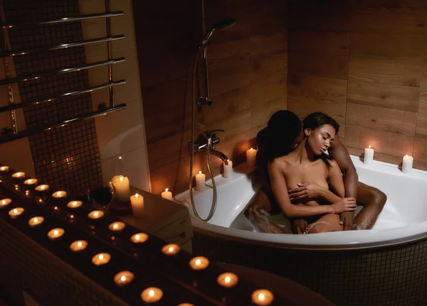 Strong desire. Young african man kissing his girlfriend in neck while taking foamy bath with candles together at home