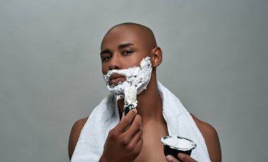 Gömleksiz, çekici, Afro-Amerikalı bir adam kameraya bakıyor, yüzüne tıraş köpüğü sürerken fırça kullanıyor, gri arka planda tek başına poz veriyor.
